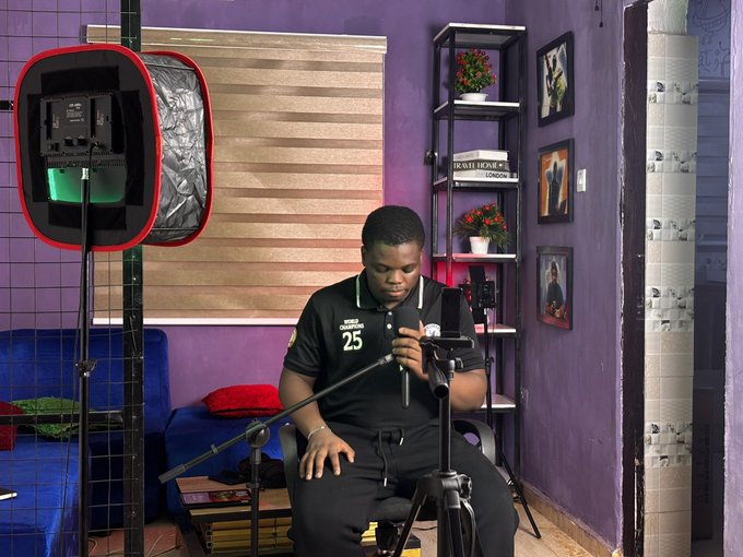A young man wearing a black polo shirt with '25' sits at a microphone setup in a purple room, with professional lighting equipment visible to his left.