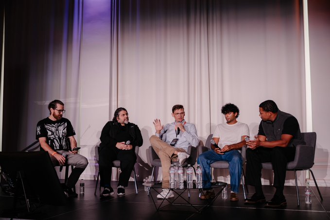 Five men sit in chairs on a stage in what appears to be a panel discussion, with water bottles on a small table in front of them.