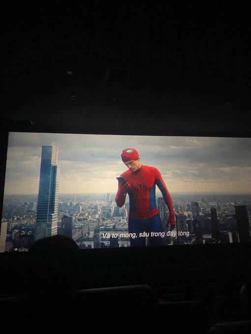 A photo of a movie theater screen showing Spider-Man looking at his phone with a city skyline behind him, with Vietnamese subtitles reading 'Và từ mong, sáu trong dạy lòng...'.