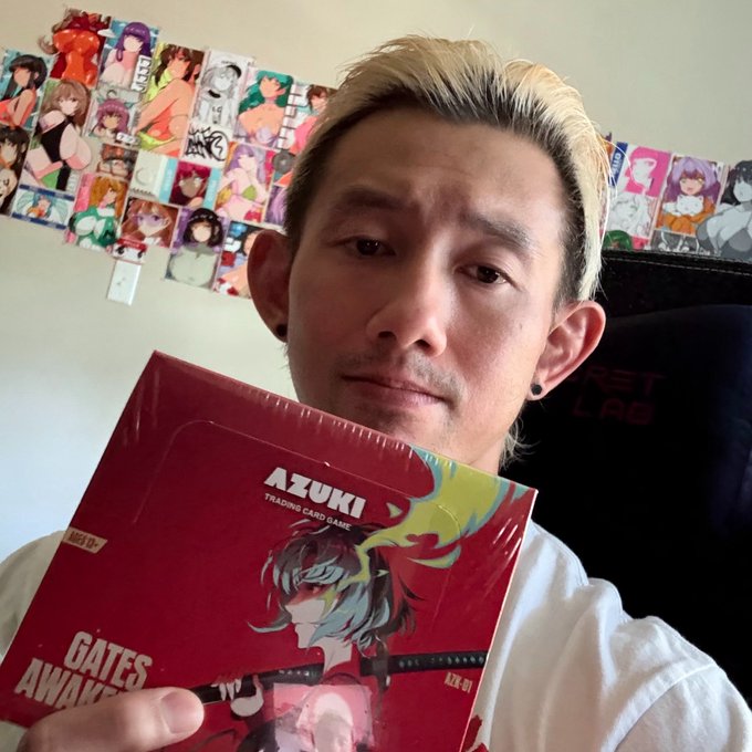 The image features a man holding up a red book with a cartoon character on the cover titled "Gates Anki". The man appears to be posing for the photo while showing off his new purchase, which seems to be an anime-themed game or card.