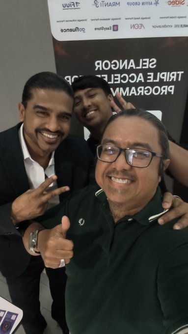 A selfie of three smiling men at what appears to be a corporate event, with a backdrop featuring sponsor logos and the text 'SELANGOR TRIPLE ACCELERATOR PROGRAM'.