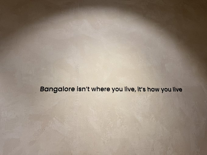 The image features a textured beige wall illuminated by a spotlight, with the text "Bangalore Isn't where you live, It's how you live" displayed prominently. The caption connects to "Indira nagar be like," humorously highlighting how the Bangalore lifestyle, particularly in areas like Indira Nagar, defines how residents experience the city rather than just their geographic location.