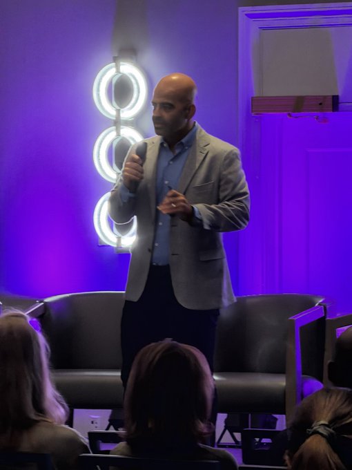 A bald man in a gray blazer and blue shirt speaks into a microphone on a stage with purple lighting and circular neon lights behind him, addressing an audience seated in the foreground.