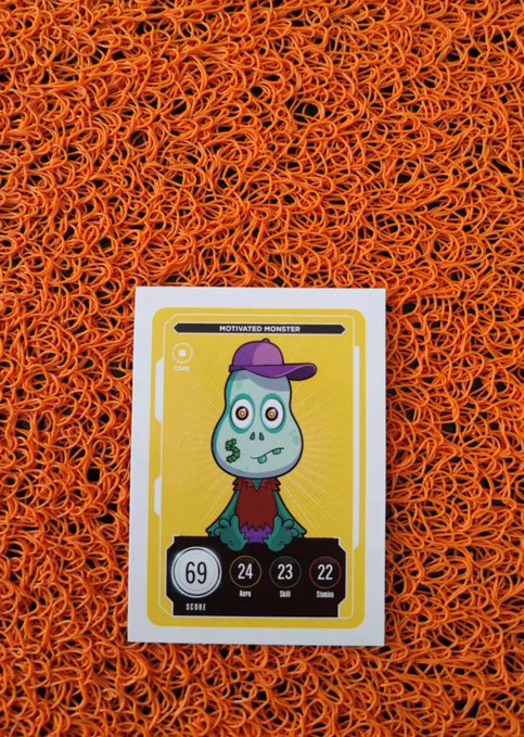 A trading card labeled 'MOTIVATED MONSTER' featuring a blue cartoon character in a purple cap rests on a textured orange mesh surface. The card displays stats: Score 69, Aura 24, Dabil 23, Stamina 22.