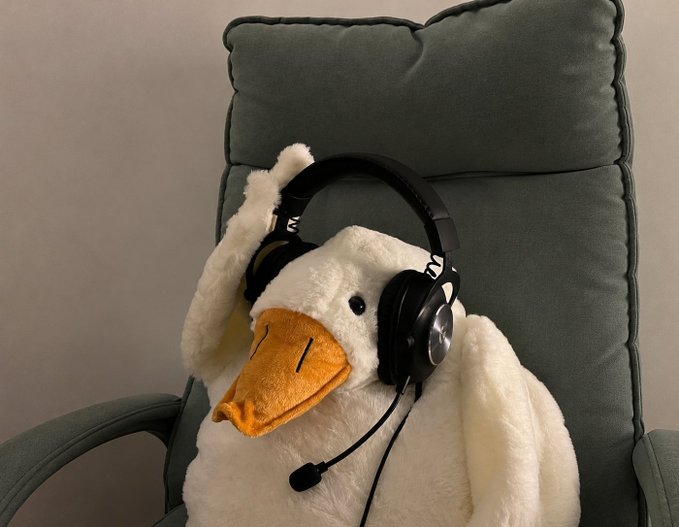 A white plush goose sitting in a green cushioned chair wearing black over-ear headphones with a boom microphone attached.