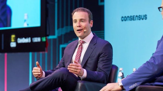 A man in a dark suit and pink striped tie speaks at a conference, sitting on a stage with 'consensus' visible in the background. Water bottles and a 'CoinDesk' display are nearby.