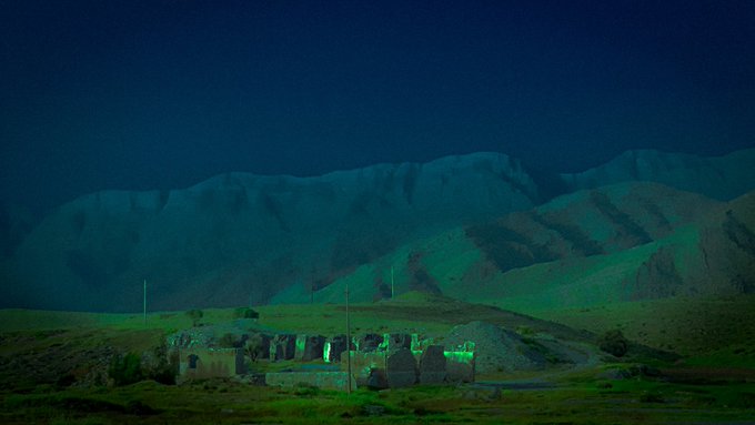 A nighttime photograph of an ancient stone village illuminated with green light against a backdrop of dark mountains.