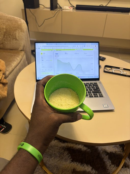 A hand wearing a green "BEST" wristband holds a green mug filled with a yellowish grainy substance (likely garri, a cassava-based food) in front of a laptop displaying a stock chart on a wooden coffee table. The image serves as visual proof of the "$GARRI" referenced in the post, suggesting the user is showcasing the staple food while reviewing financial or trading data in the background.