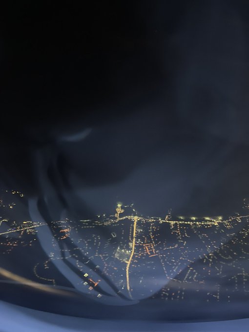 An aerial nighttime view of a city with illuminated streets and a prominent lit tower, viewed through what appears to be a scratched or distorted window or lens.