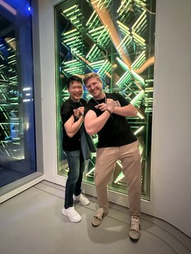 Two smiling men pose together in front of a wall featuring stacked physical Binance merchandise boxes with green geometric lighting, celebrating Binance and BNB Chain's 9th anniversary as referenced in the post.