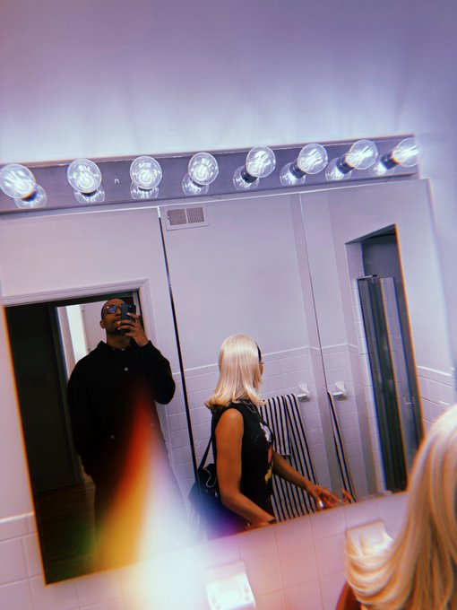 A man in sunglasses takes a mirror selfie with a camera while a blonde woman in a black top stands beside him in a bathroom illuminated by Hollywood-style vanity lights. The image ties into the post's theme of confidence, visually reinforcing the boastful swagger implied by holding $ANSEM.