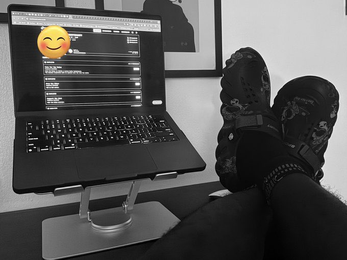 A black-and-white photo showing a person's feet in Merrell water shoes propped up on a desk next to a MacBook displaying a web page with a smiley face emoji covering the screen.