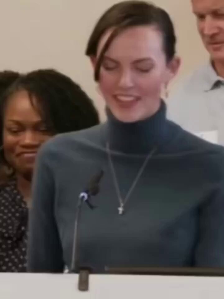 The image shows a woman wearing a gray turtleneck sweater, standing at a podium with her hands behind her back as she speaks into a microphone. She appears to be delivering a speech or presentation. The background of the image features two other people, one on each side of the speaker, suggesting that they are also present and listening to what she has to say.