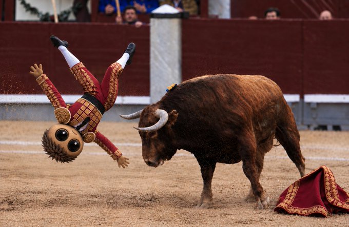 A surreal and humorous scene in a bullring shows a matador character costume head being launched upside-down into the air, as a large brown bull charges past a discarded red and gold cape on the sandy ground.