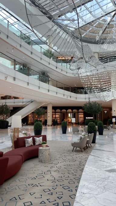 The image shows the elegant atrium of an upscale shopping mall featuring a grand staircase, a large cascading crystal chandelier, plush burgundy seating, and polished marble floors. Visible storefronts include "DEAN & DELUCA" and "BURBERRY," aligning with the "GM" (general mall) reference by highlighting the upscale retail environment.