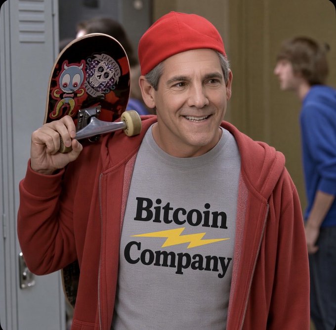 A man wears a red beanie and red hoodie over a gray t-shirt reading 'Bitcoin Company' with a lightning bolt, holding a skateboard on his shoulder in what appears to be a school hallway.