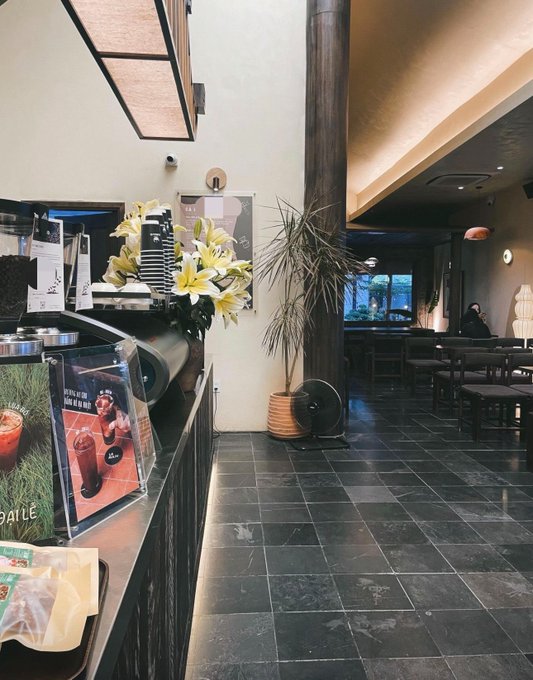 The interior of a modern café with a coffee counter, lilies in a vase, potted plant, and dining area in the background.