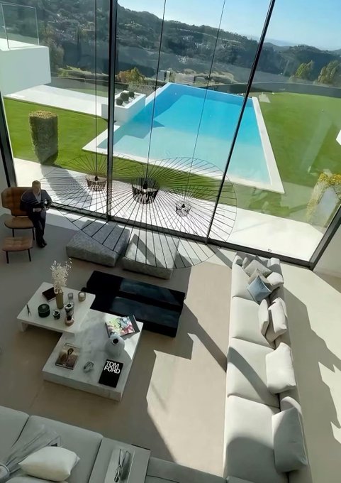 An aerial view of a modern luxury home interior with white sofas, a coffee table with a Tom Ford book, and floor-to-ceiling windows overlooking a blue swimming pool and hillside landscape.