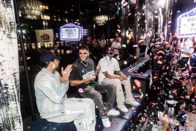 Two men sit at a venue surrounded by falling colorful confetti, with one wearing a New York Yankees cap and the other in a black shirt and sunglasses, while a 'WELCOME' sign is visible in the background.