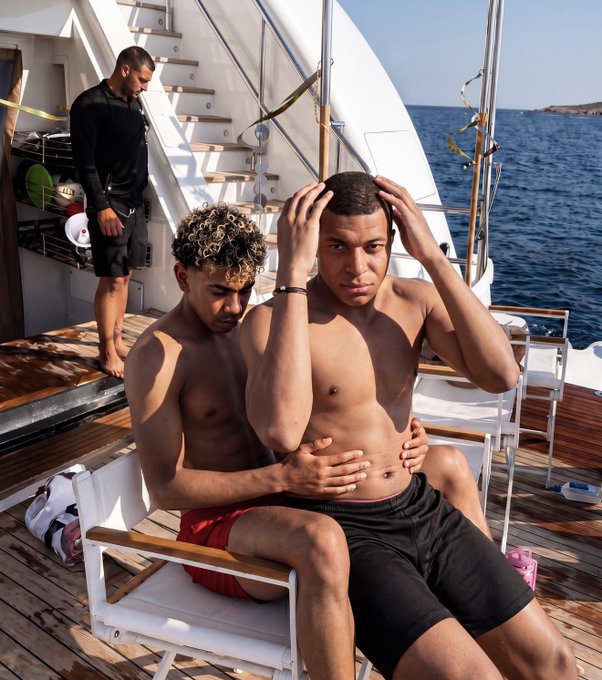 Two shirtless young men sit closely together on a wooden lounge chair aboard a sailboat, with one holding his hands on his head while the other embraces him from behind, as a third person stands nearby against the backdrop of open blue sea—a sensual, summery scene fitting the aesthetic of a luxury resort or yacht lifestyle hashtag.