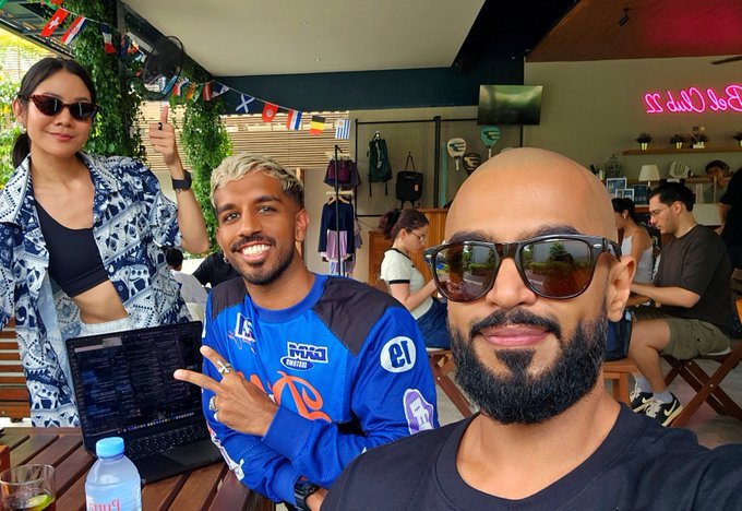 A selfie taken at an outdoor café featuring three people—a woman with dark hair and sunglasses in a blue patterned jacket, a man with bleached blonde hair in a blue esports jersey, and a bearded man with sunglasses in the foreground—posing together near a laptop.