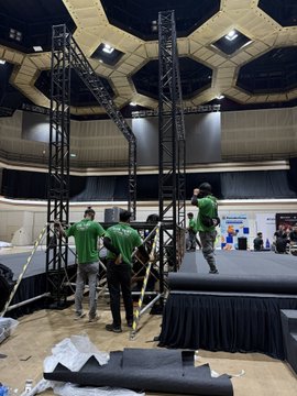 Several workers in green shirts assemble a black metal truss structure on a stage inside a large hall with a distinctive hexagonal honeycomb ceiling pattern.