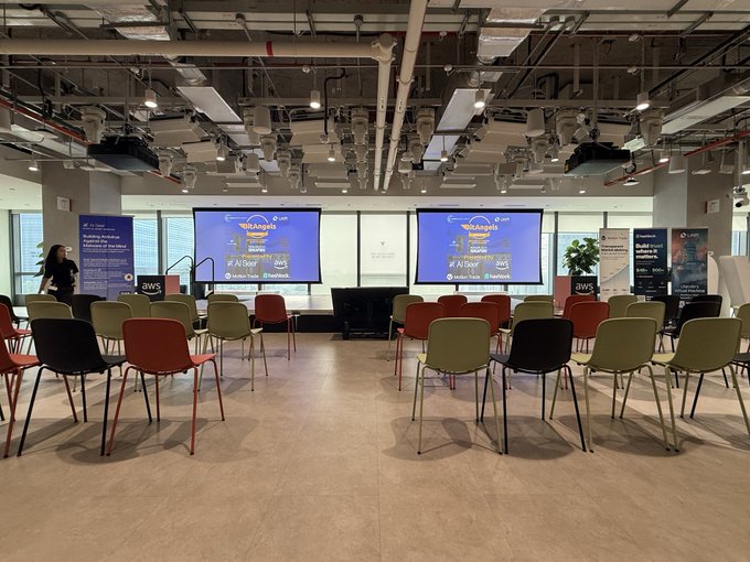 An empty event space is set up with rows of green and red chairs facing two screens displaying 'SãoAngels' and '#AngelSummit 2025' graphics, with a stage area featuring AWS banners.