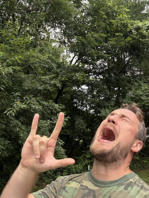 A selfie of a bearded man in a camouflage t-shirt making a 'rock on' hand gesture with his mouth wide open, posed against a background of dense green forest trees.