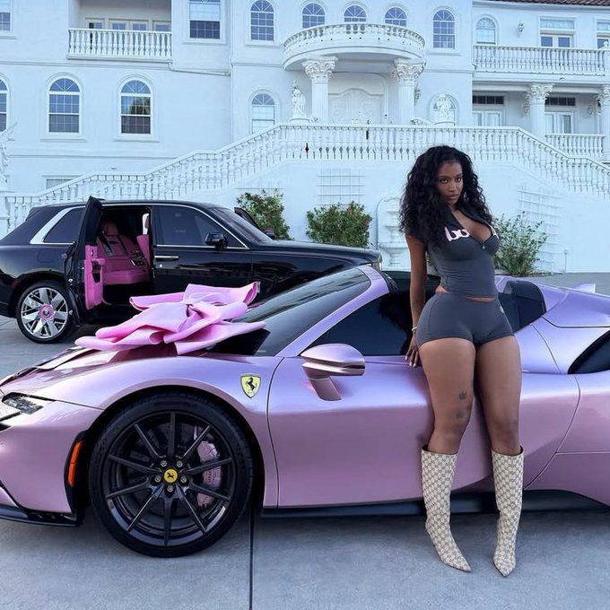 A woman in a dark outfit and white boots standing next to a pink/lavender Ferrari with a large pink bow on the hood, with a black SUV parked in front of a mansion.
