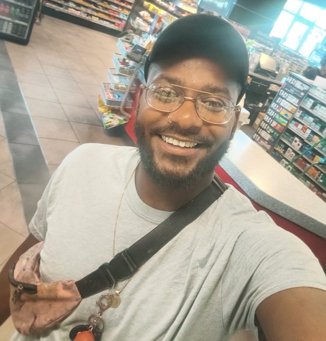A smiling man wearing a black cap, clear glasses, and a gray t-shirt takes a selfie inside what appears to be a convenience store or gas station, with shelves of products visible behind him.