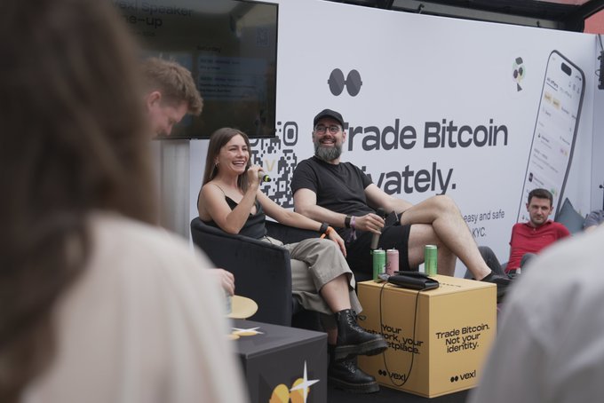 The image shows a group of people gathered together at an event, with some sitting down and others standing up. They are engaged in conversations about Bitcoin and its impact on society. The attendees include both men and women, all dressed casually. There is a large screen displaying information related to Bitcoin, which serves as the backdrop for their discussions. One person can be seen holding a cell phone, possibly taking notes or sharing information with others at the event.