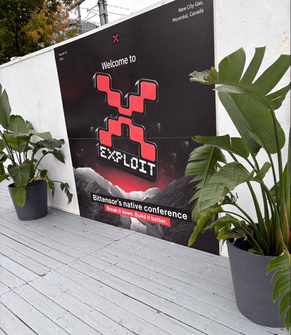 A large welcome sign for 'VS EXPLOIT,' Bittensor's native conference in New City Gas, Montréal, Canada, flanked by large potted plants on a wooden deck.