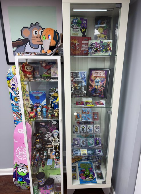 A glass display cabinet showcasing various vinyl figures, trading cards, and collectibles, with a 'Worlds Apart' skateboard and pixel art hanging beside it.