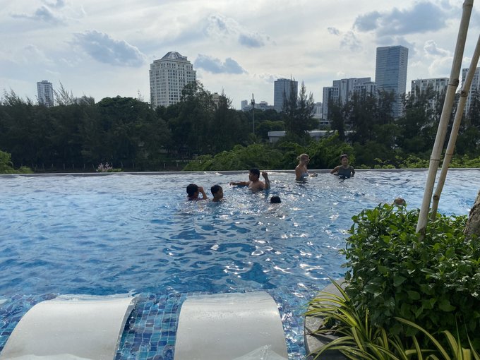 Children and adults swimming in an infinity pool overlooking a modern city skyline with tall buildings and green trees under a partly cloudy sky.