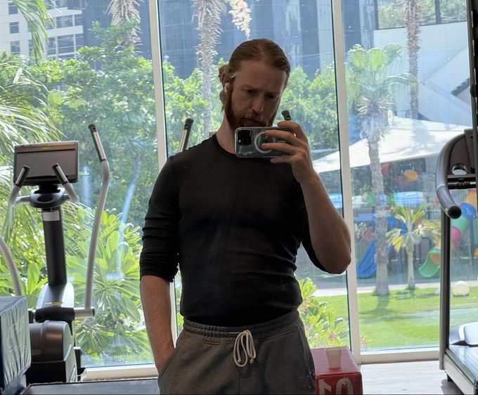 A man with a beard and ponytail takes a mirror selfie in a gym, wearing a black long-sleeve shirt and gray sweatpants, with palm trees and a children's play area visible through the window.
