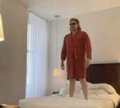 A low-resolution, slightly blurry photo shows a man wearing a red outfit standing in a bedroom between a bed and white curtains.