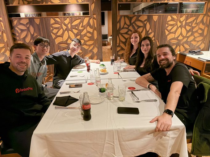 Group of six people smiling around a white-clothed restaurant table with Coca-Cola bottles and table settings, including a man in a 'RedotPay' hoodie on the left.