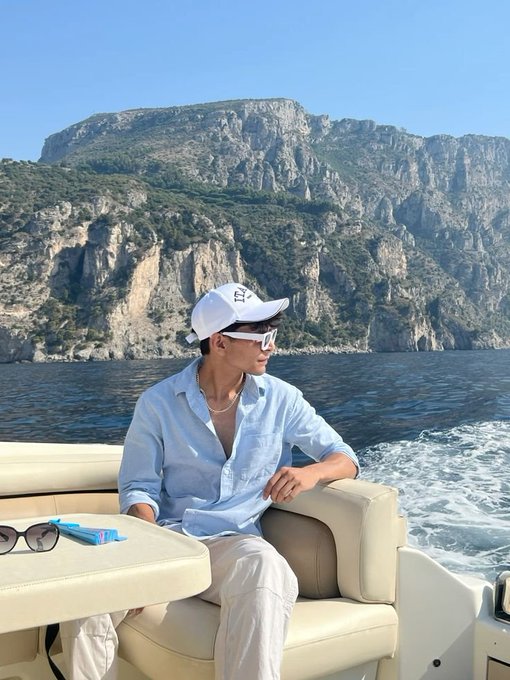 A stylish young man wearing a light blue shirt, white cap, and sunglasses sitting on a boat with a dramatic rocky coastline and sea in the background.