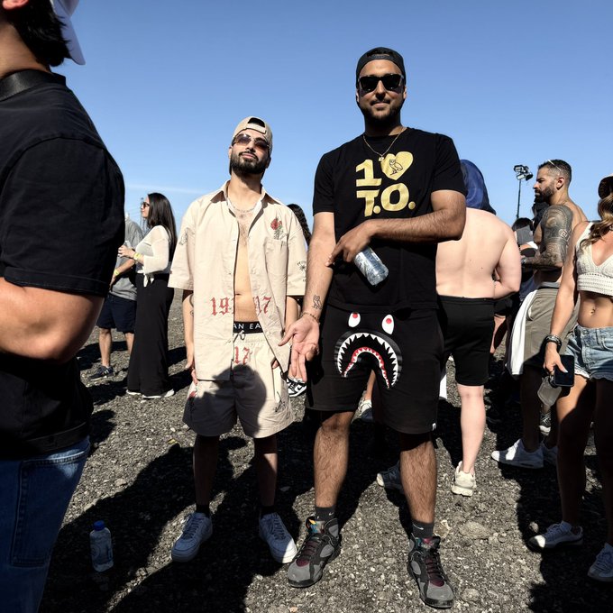 Two stylish young men pose at an outdoor event, with the man on the right wearing a black "1/10" t-shirt and shark-print shorts, while his companion sports a cream-colored button-up featuring "19__17" and rose graphics—both outfits reflecting the bold streetwear aesthetic that aligns with ANSEM's 10bil+ milestone celebration.