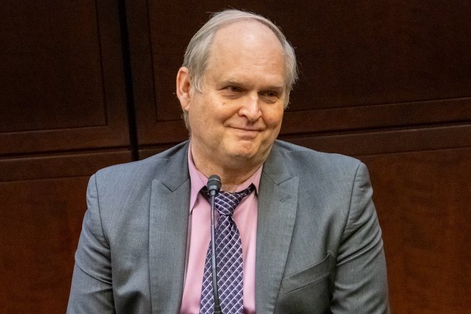 A photograph of a smiling older man in a gray suit, pink shirt, and patterned tie sitting at what appears to be a hearing or panel with a microphone in front of a wood-paneled background.