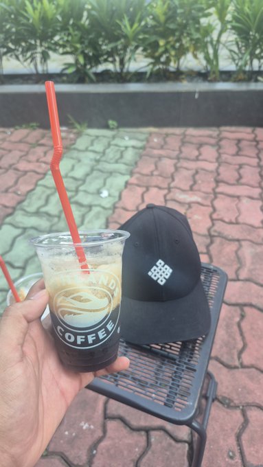 A person holds an iced coffee in a clear plastic cup with "BLEND COFFEE" branding and a red straw, with a black cap beside it on a chair. The image ties into the post's ritual theme of coffee and lifestyle, reinforcing the relaxed, daily-habit aesthetic the $RITUAL post conveys.