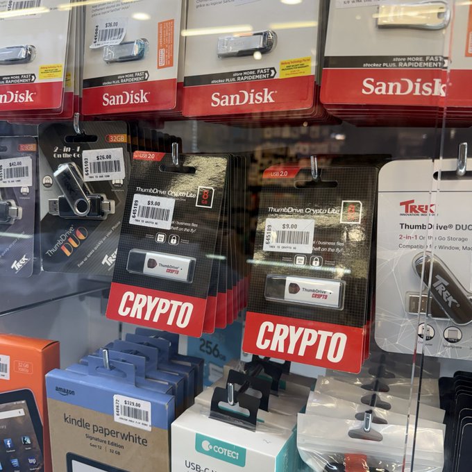 A display case at Mustafa Centre in Singapore's Little India features Trezor and Ellipal hardware wallets labeled "CRYPTO" alongside everyday electronics like SanDisk USB drives and a Kindle Paperwhite, with the Trezor devices priced around $9.99–$13.99. The image humorously illustrates the post's point about crypto going mainstream, as these specialized digital asset storage devices sit on store shelves right next to ordinary consumer tech.