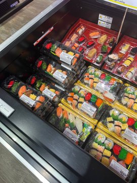 The image shows a display case filled with various types of sushi, including both vegetarian and non-vegetarian options. The sushi comes in different shapes and sizes, making for an enticing selection for customers. The SuperCashMe campaign encourages shoppers to take advantage of their cashback offer by recording a QR code payment video on social media platforms or entering the code provided in the image. This allows them to receive a full refund of their transaction value if they can prove that they made the purchase through SuperCashMe's website.