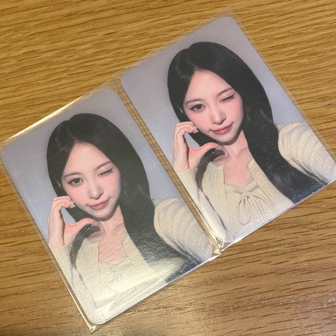 Two photocards featuring the same portrait of a young woman with long dark hair, wearing a cream-colored sweater, displayed inside clear protective sleeves on a wooden surface—unrelated collectibles that humorously contrast with the post about becoming a Ripple cryptocurrency holder.