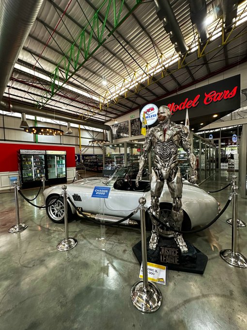 A silver chrome robotic statue stands next to a classic silver sports car with 'CHASING 2000' on its license plate, displayed inside a 'Model Cars' showroom with green-trussed ceiling.