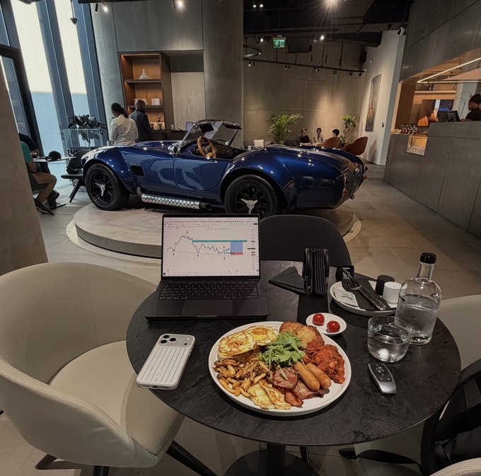 A laptop displaying a "S&P 500" chart sits on a table alongside a hearty breakfast plate and car keys, with a blue Shelby Cobra sports car displayed in the background of a modern showroom—capturing a lifestyle moment that ties the "gm ct" market post to aspirations of financial success and luxury rewards.