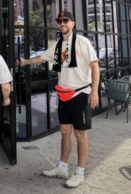 A man standing on a city sidewalk wearing a white t-shirt, black shorts, an orange fanny pack, a baseball cap with an 'S' logo, and sunglasses, with a black and white towel draped around his neck.