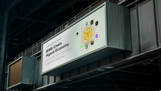 An illuminated billboard in a large industrial space advertising 'BNB Chain Agent Economy' with various cryptocurrency logos around a central golden emblem.