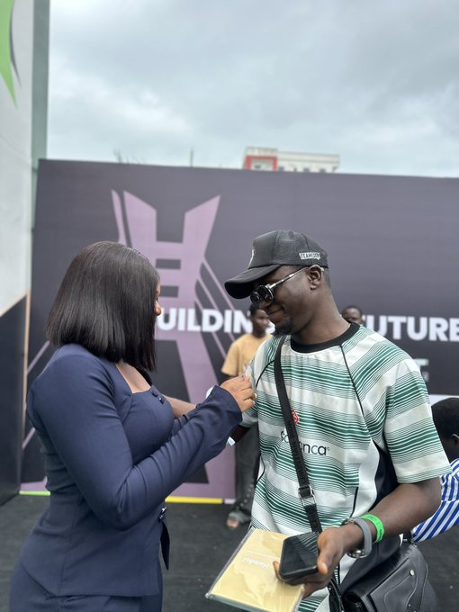 A woman in a dark blue top interviews a man wearing a black cap, sunglasses, and a striped 'Agbenc' shirt at an outdoor event with a banner reading 'BUILDING' and 'FUTURE' in the background.