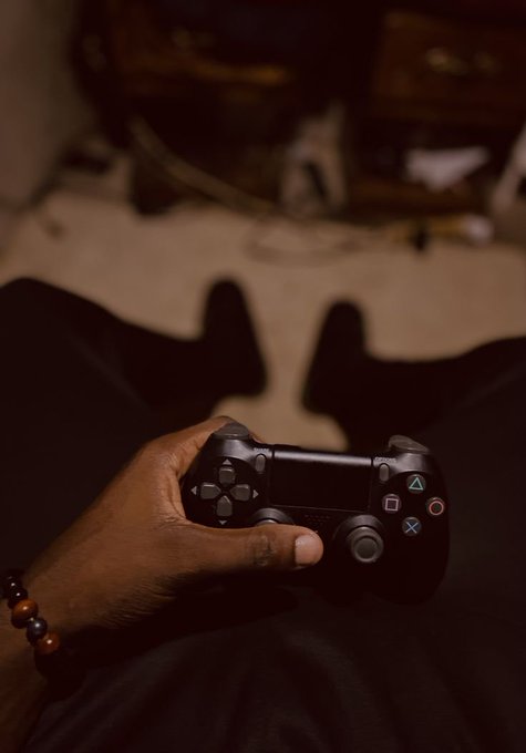 A person's hand holding a black PlayStation 4 DualShock controller in a dimly lit room, with the person wearing a beaded bracelet on their wrist, sitting with legs stretched out.